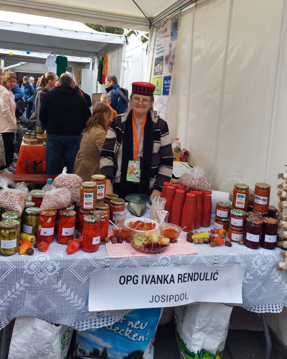 Baka Ivanka za štandom OPG-a Ivanka Rendulić na sajmu, okružena teglama zimnice i pletenicama češnjaka